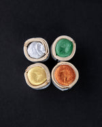 4 shimmery Beam Paints water colour paintstones in silver, green, gold, and red. The watercolours are wrapped in waxed canvas and are sitting on a black background.