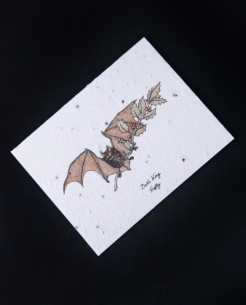 Cream-coloured card with illustration of a bat holding a branch of holly. The paper is naturally textured and studded with seeds.