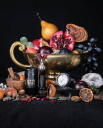 Items from LVNEA's Feast of Fools gift box, including a black glass jar of incense, a black glass bottle of eau de parfum, an open black glass jar of body butter, and a silver pomander charm hanging from a red silk ribbon. The items are surrounded by a still-life of fruit cascading out of a brass bowl, and dried fruits and spices strewn about a black linen surface.