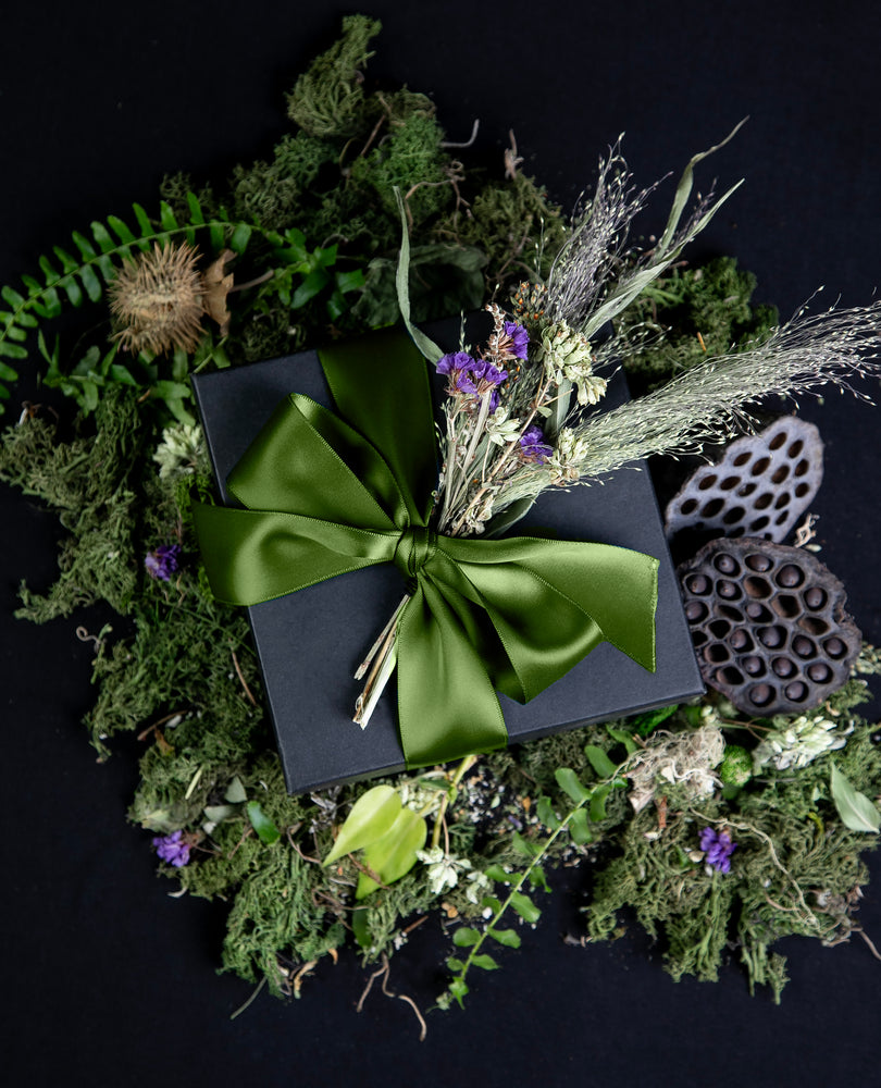 Black cardboard box wrapped in an olive green silk ribbon, sitting atop lush greenery and botanicals. The box contains LVNEA's limited edition THE WITCH'S GARDEN seasonal offerings.