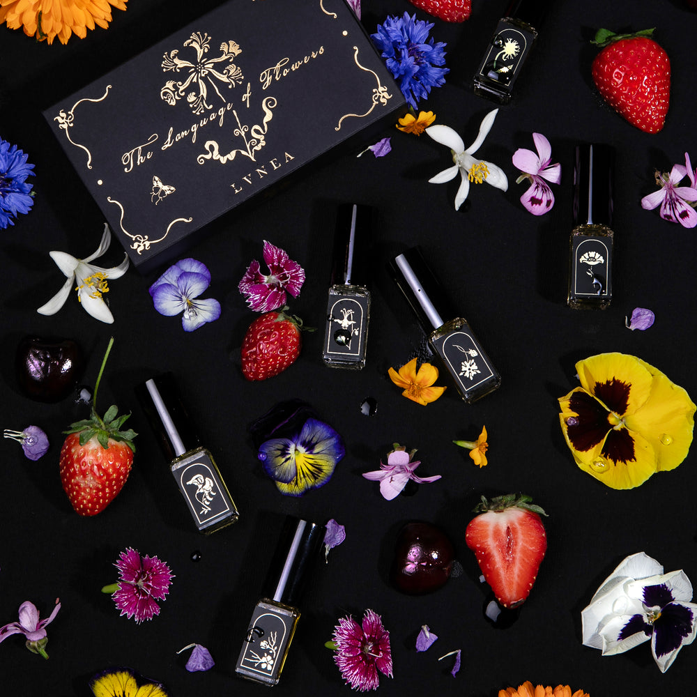 Overhead shot of all 6 "Language of Flowers" vials seen from overhead, scattered amongst fresh flowers next to their closed box.