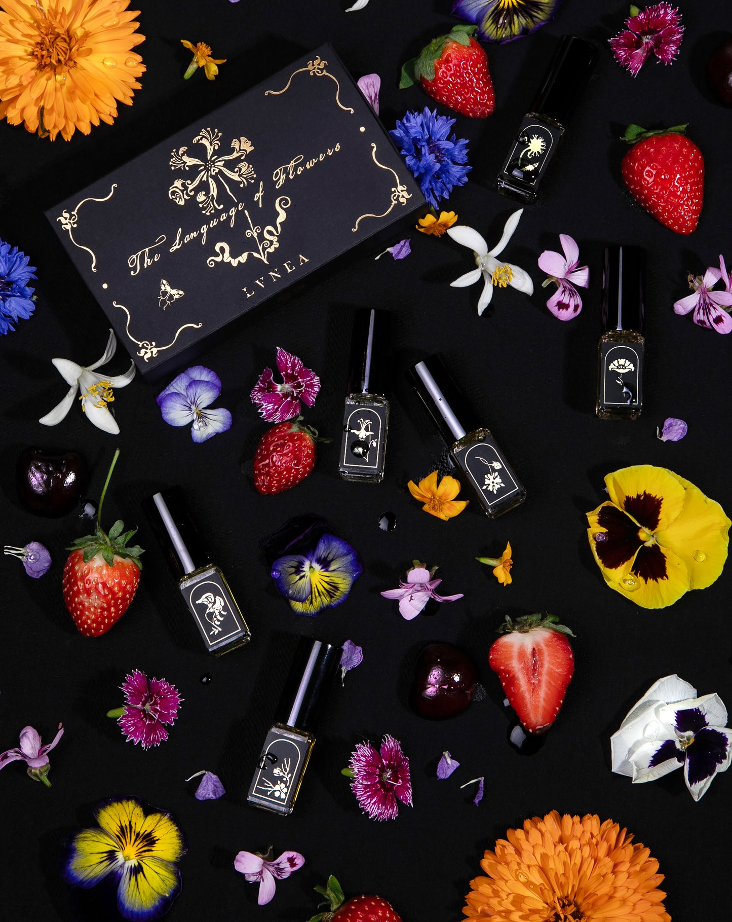 Overhead shot of all 6 "Language of Flowers" vials seen from overhead, scattered amongst fresh flowers next to their closed box.