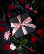 Black box tied with a light pink silk ribbon, sitting on black velvet fabric, with scattered rose petals and full rose stems surrounding it.