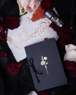 Overhead shot of a hand-bound scented paper booklet and black bottle of handmade ink, surrounded by rose petals. There is a white paper sheet with the  word "Devotion" scrawled in cursive with red ink.