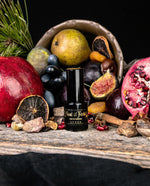 15ml black glass bottle of LVNEA's Feast of Fools Eau de Parfum surrounded by a cornucopia of fresh and dried fruit, including pomegranates, grapes, pears, orange slices, cinnamon sticks, figs, and other edible goodies.
