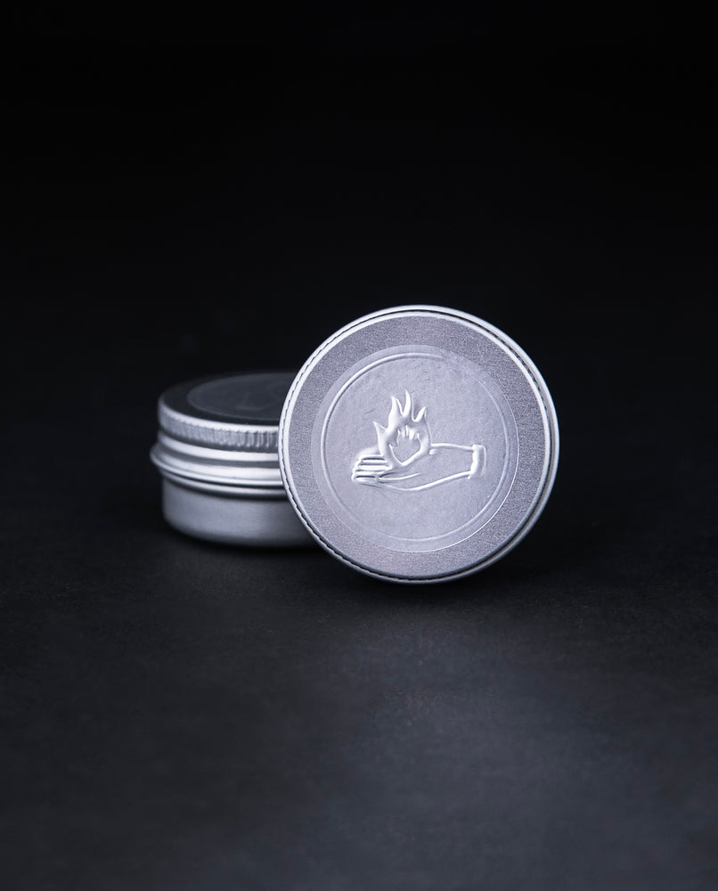 2g sample of LVNEA's "Fire and Oud" solid perfume presented in a metal tin. The lid is embossed with an illustration of a hand holding a flame.