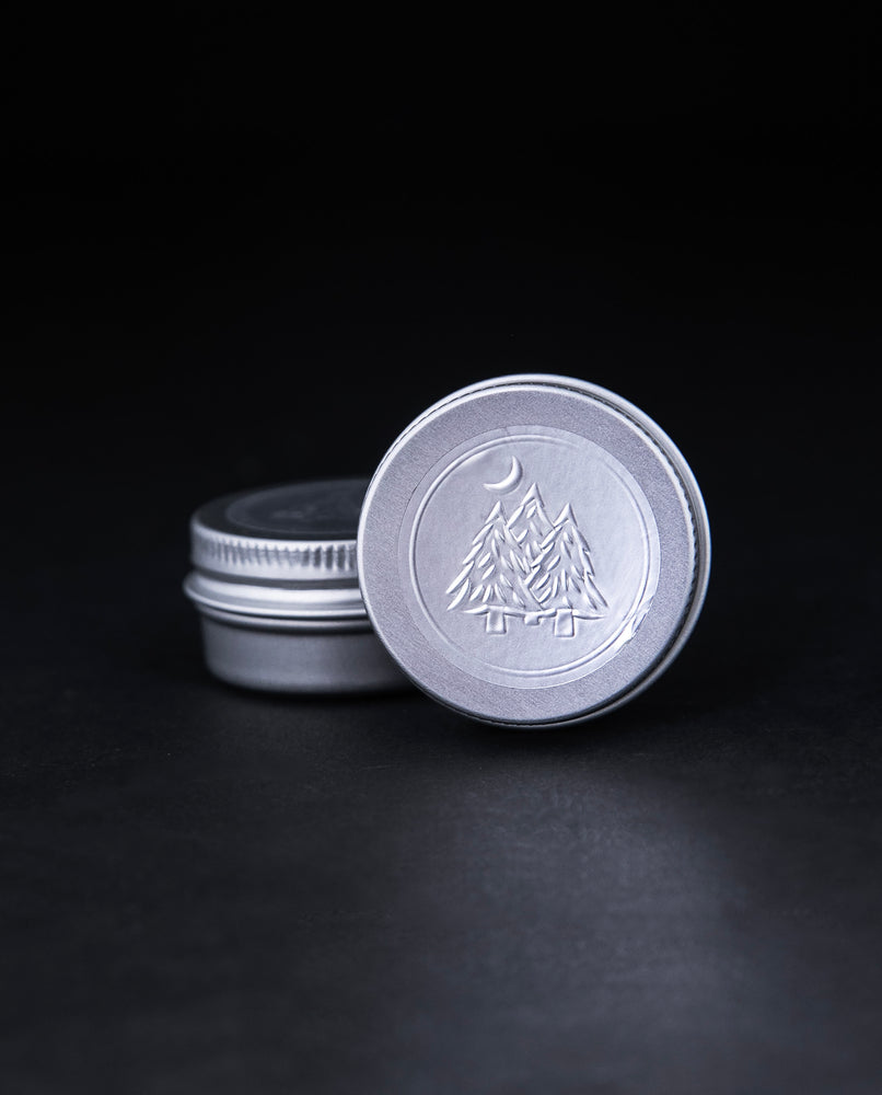 2g sample of LVNEA's "Hemlock Shade" solid perfume presented in a metal tin. The lid is embossed with an illustration of 3 pine trees with a crescent moon overtop.