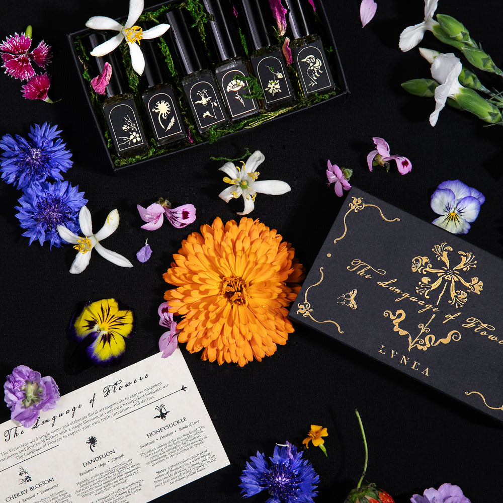 
                      
                        "Language of Flowers" box set, open to reveal 6 mini vials of eau de parfum nestled together in a bed of lush green moss and rose petals. It is surrounded by the box lid, box insert, and loose colourful botanicals. 
                      
                    