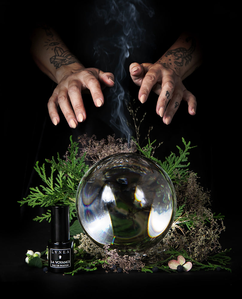 Two hands emerge from the darkness over a crystal ball surrounded by a plume of smoke, moss and juniper. A 15ml black glass bottle of LVNEA's "La Voyance" natural perfum sits at the base of the crystal ball.
