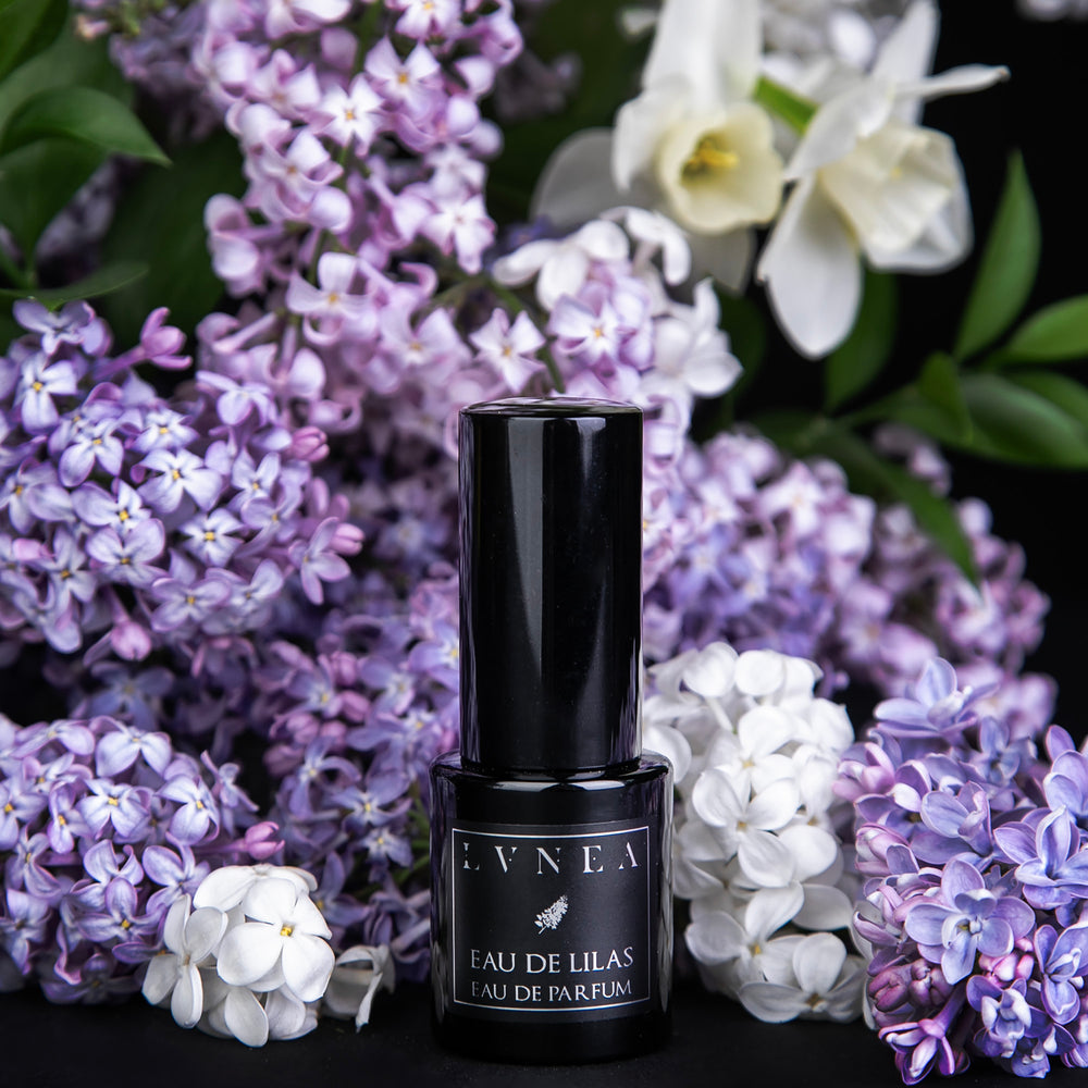 15 ml black glass bottle of LVNEA's limited edition Eau de Lilas perfume, surrounded by fresh lilac blossoms and other florals on black background