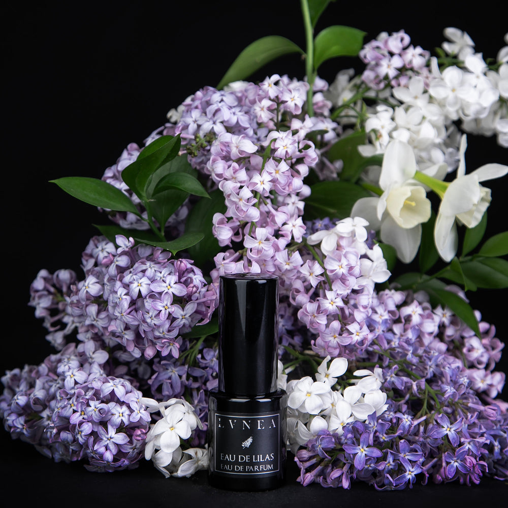 15 ml black glass bottle of LVNEA's limited edition Eau de Lilas perfume, surrounded by fresh lilac blossoms and other florals on black background