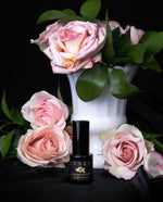 15 ml black glass bottle of LVNEA's "Rose Devotion" Limited edition eau de parfum, stamped with gold foil. The bottle is sitting on black silk, and surrounded by fresh pink roses, one of which is dripping with honey.