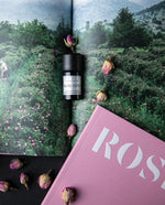 bottle of rose otto essential oil sitting atop an open book showcasing a photo of a rose field
