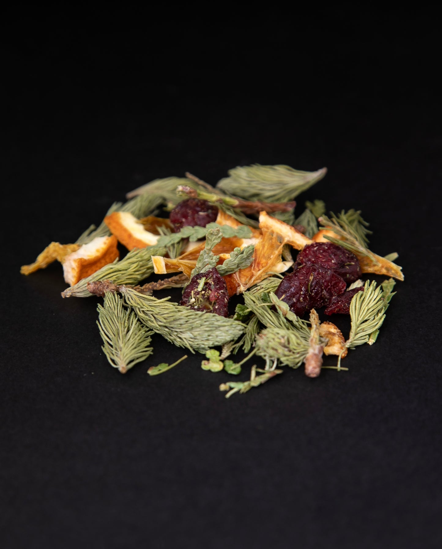 a handful of loose lead "Solstice" herbal tea against a black background. This colourful blend features dried cranberries, spruce tips, and dried orange slices.