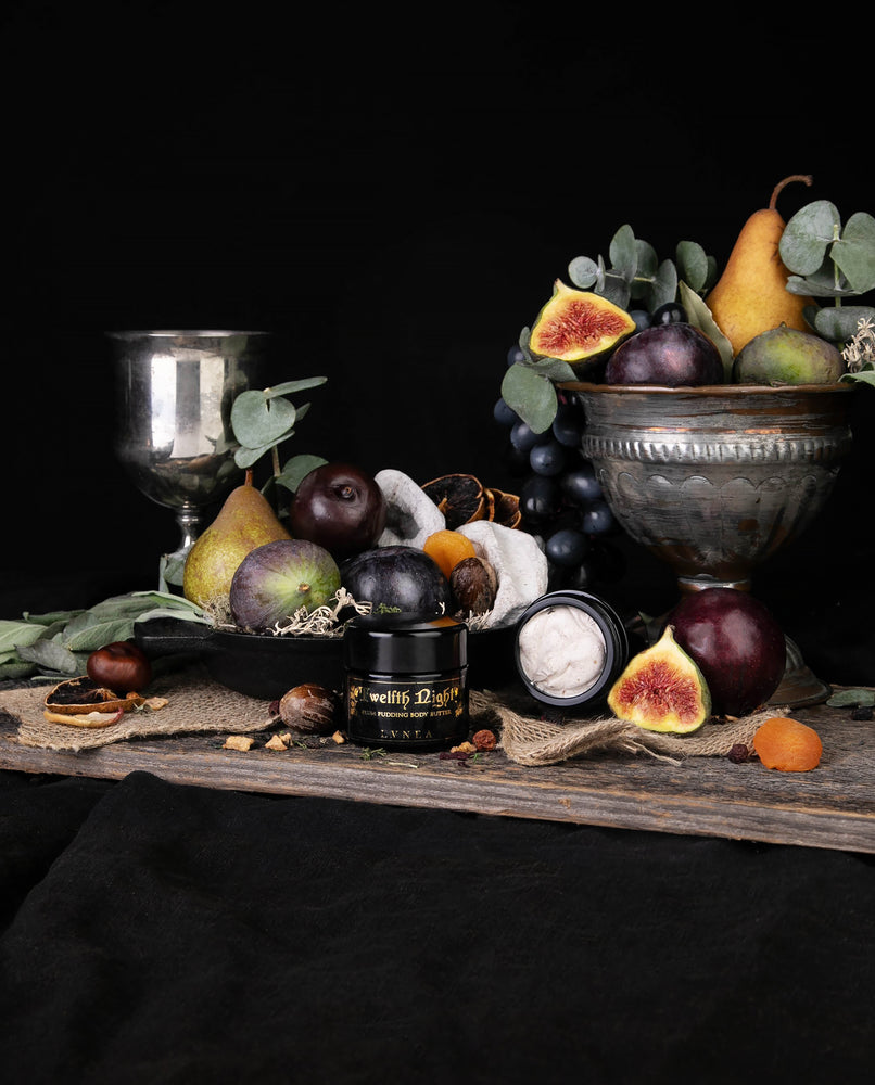 Black glass jar of LVNEA's Twelfth Night body butter surrounded by figs, plums, and other fruits.