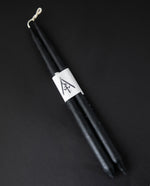 A pair of black beeswax taper candles on a black background. They are bound together by a narrow paper label printed with RIITUAALIA'S logo.