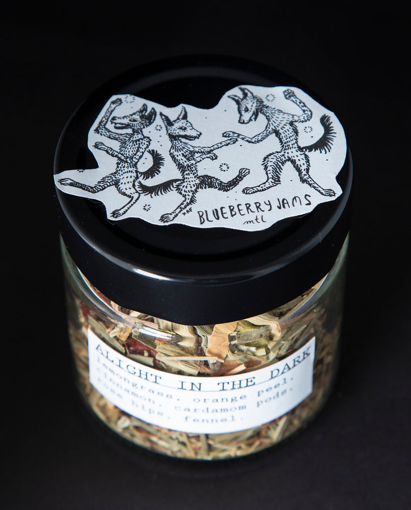3/4 view of a clear glass jar with black screw on top containing blueberryjams' "Alight In The Dark" herbal tea blend. There is an illustration of foxes on the top of the jar's lid.