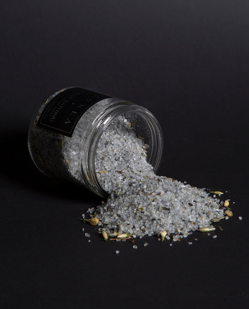 LVNEA'S Lunar Bathing bath salts spilling out of a tipped over 16 ounce clear glass jar. Milky oat tops, jasmine blossoms, and lavender buds are visible in the salt blend.