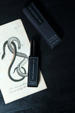 30ml bottle of La Serpentine and its box laying atop a black wooden surface and an antique illustration of two entwined serpents