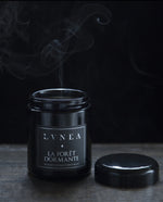 La Foret Dormante candle in an 8oz black jar with lid removed. A plume of smoke is coming out from the top of the candle.