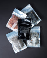 4 ounce black resealable pouches of all 5 of LVNEA's ritual bath salts. Includes: Fire Bathing, River Bathing, Ocean Bathing, Forest Bathing, and Lunar Bathing lying flat on a black background