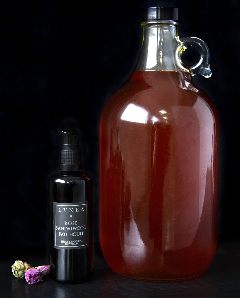 Black bottle of LVNEA's Rose, Sandalwood and Patchouli body serum standing next to large clear glass jug filled with amber-coloured oil
