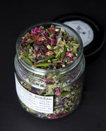 Clear glass jar with lid open, revealing a tea blend containing labrador tea, rose petals, and other herbs.