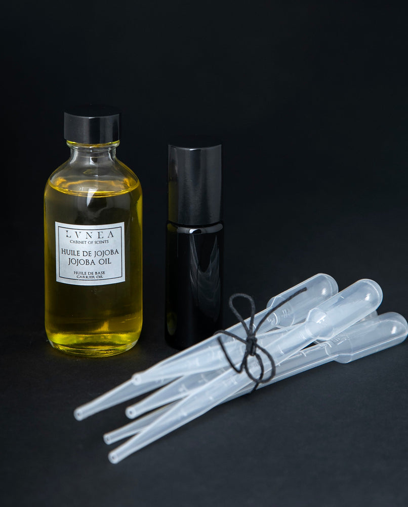 Clear 60ml glass bottle of golden jojoba oil, a 10ml black glass roller, and 5 clear plastic pipettes bundled together with a piece of black twine.