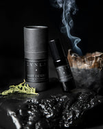 10ml bottle of LVNEA'S Nuit Désert perfume leaning up against its black packaging. They are propped up on a black piece of wood. There is a plume of smoke in the background and a piece of cedar leaf in the foreground.