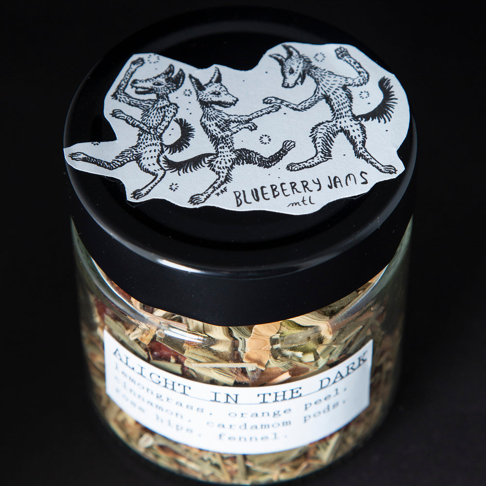 3/4 view of a clear glass jar with black screw on top containing blueberryjams' "Alight In The Dark" herbal tea blend. There is an illustration of foxes on the top of the jar's lid.
