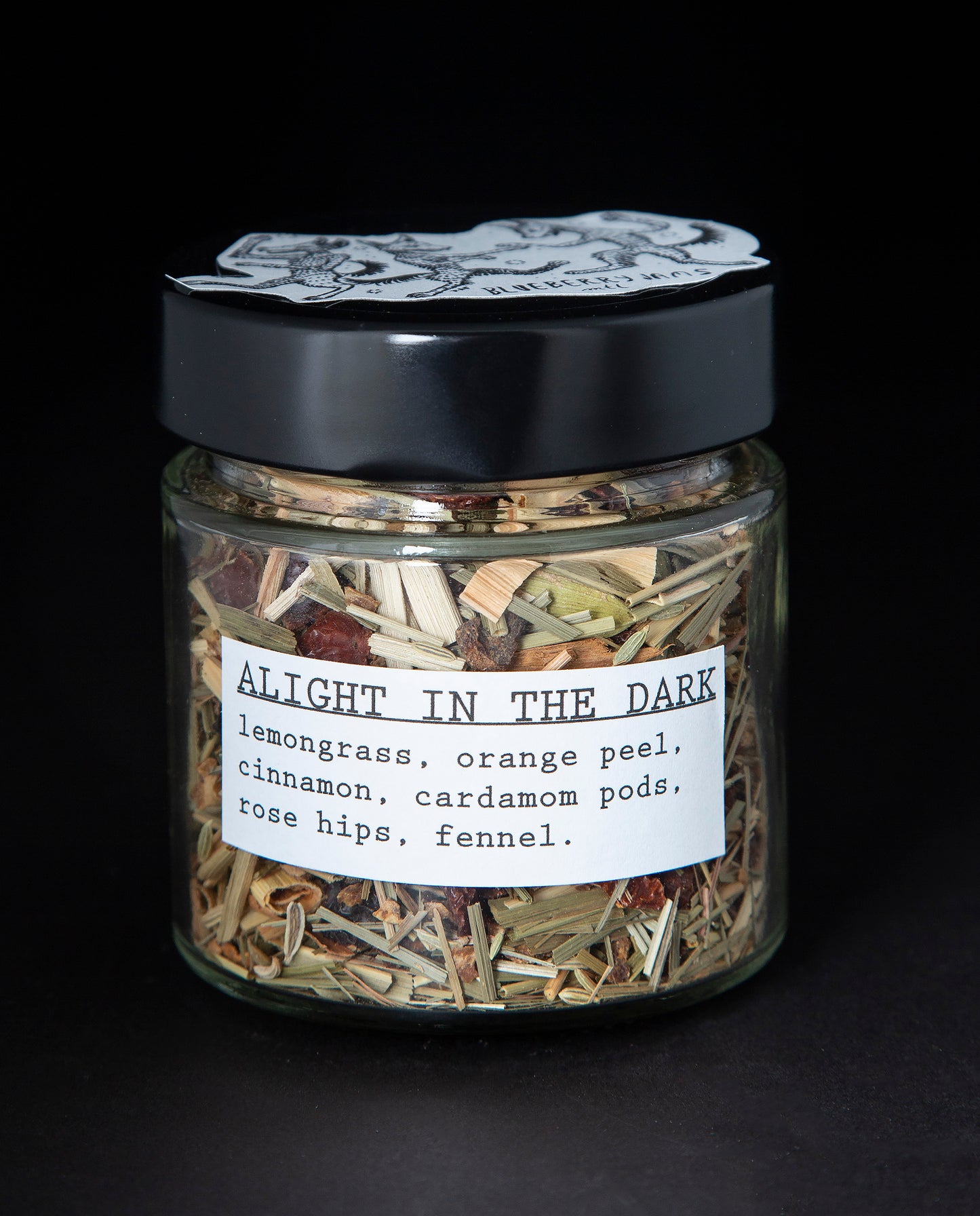 Clear glass jar with black screw on top containing blueberryjams' "Alight In The Dark" herbal tea blend. Presented on black background.
