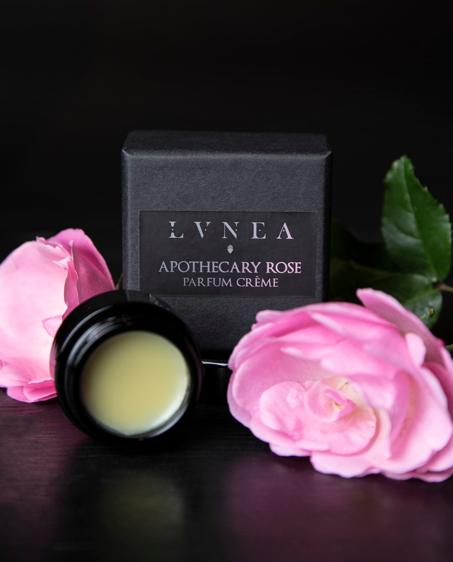Opened black glass jar of Apothecary Rose solid perfume and black box next to roses on a black wood surface