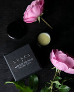Opened black glass jar of LVNEA'S Apothecary Rose solid perfume and black box next to roses on a black wood surface 
