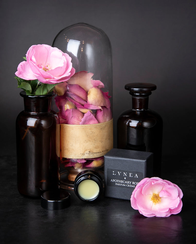 10g jar of solid perfume next to fresh roses and apothecary jars on black surface