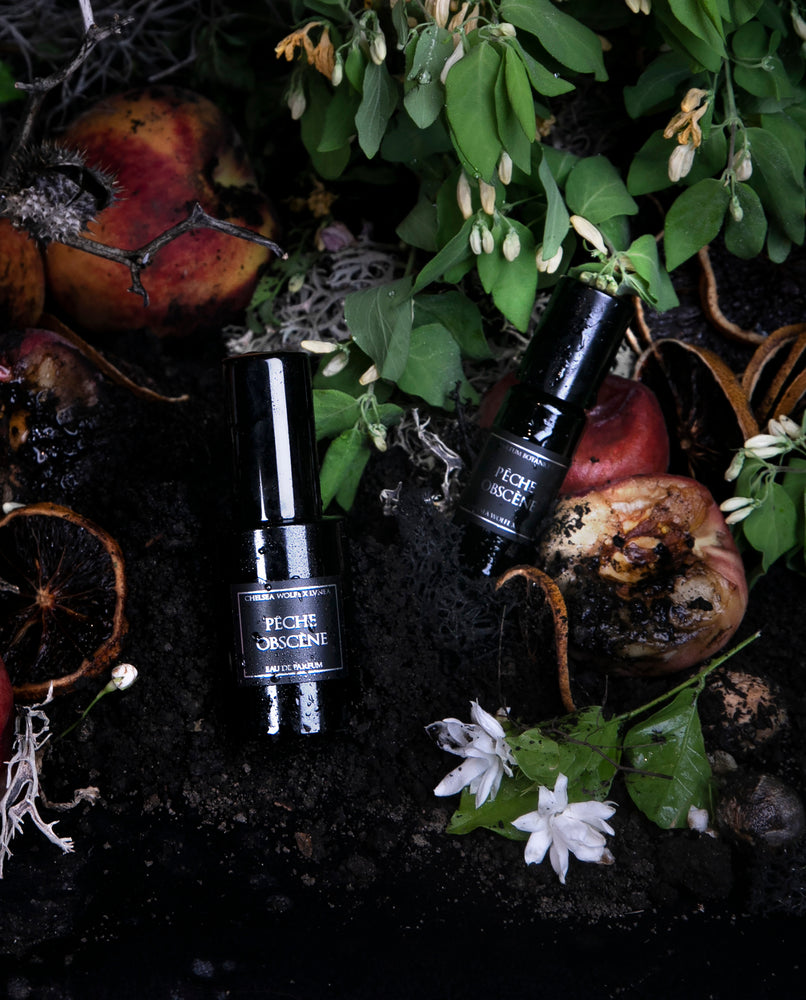 30ml and 10ml bottles of LVNEA's best-selling Pêche Obscène natural fragrance lying in dirt surrounded by peaches and botanicals.