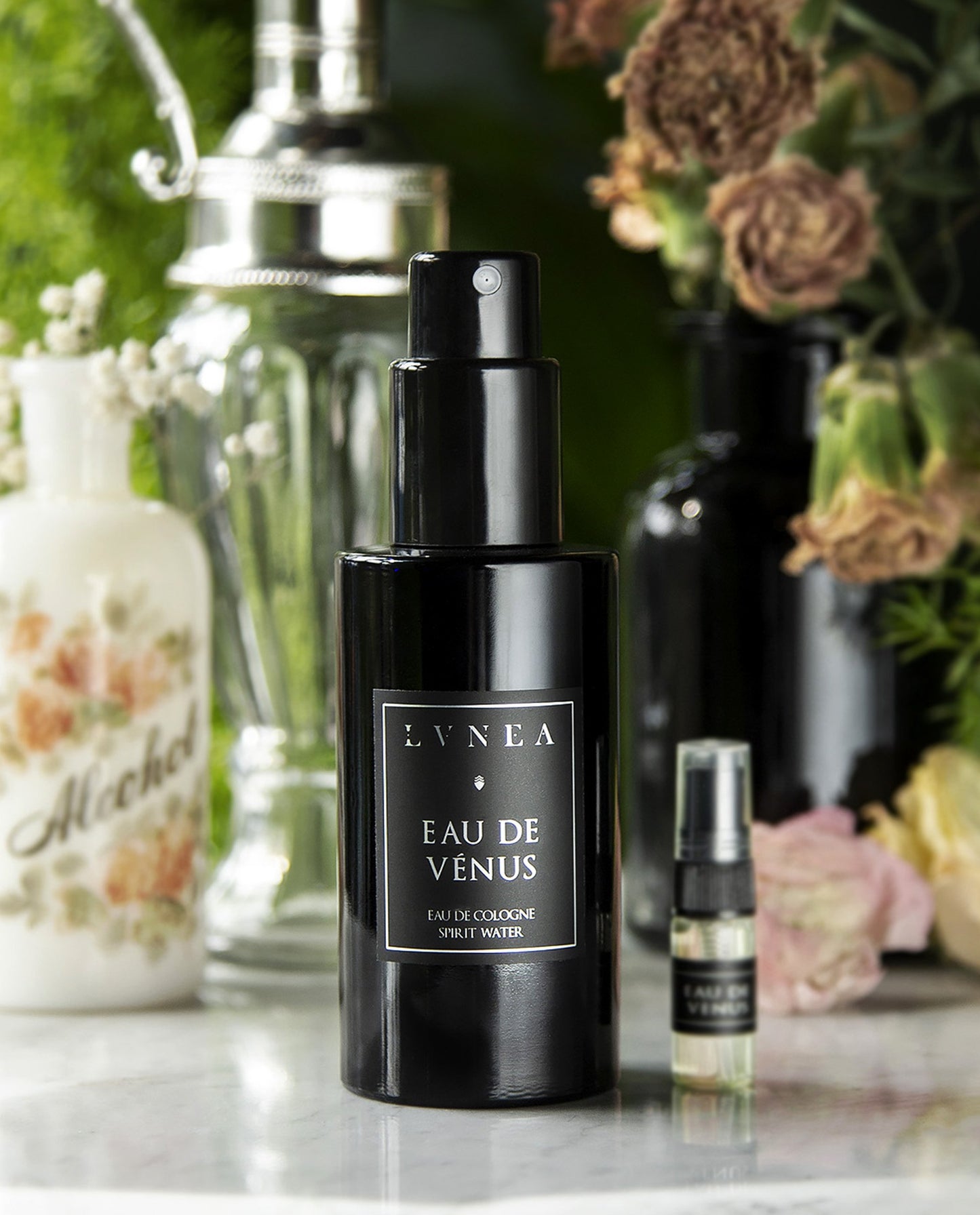 100ml black bottle and sample vial of LVNEA'S Eau de Venus fragrance sitting on white marble surrounded by pink florals