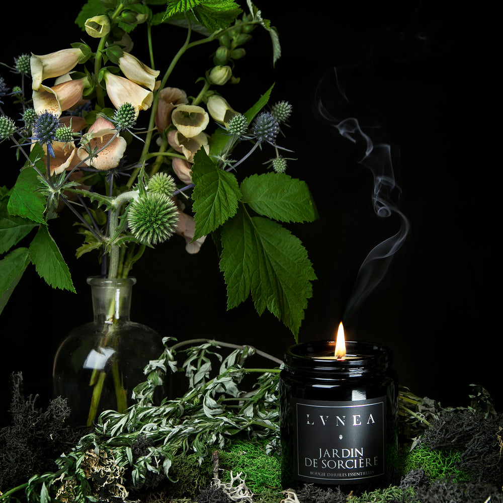 A lit Jardin de Sorcière candle displayed on a bed of moss, with a bouquet of native garden flowers in the background.