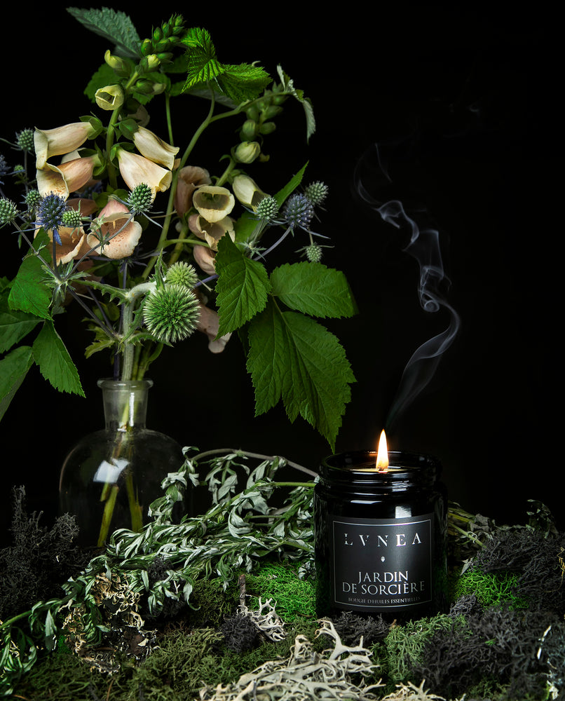 A lit Jardin de Sorcière candle displayed on a bed of moss, with a bouquet of native garden flowers in the background.
