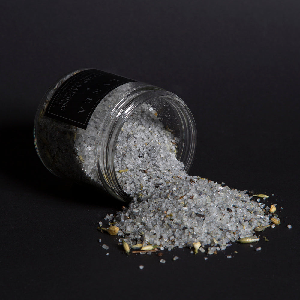 LVNEA'S Lunar Bathing bath salts spilling out of a tipped over 16 ounce clear glass jar. Milky oat tops, jasmine blossoms, and lavender buds are visible in the salt blend.