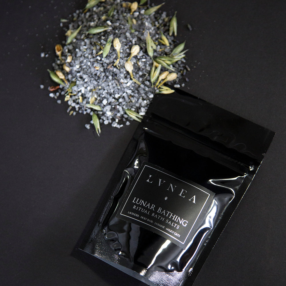 4oz black resealable pouch of LVNEA's Lunar Bathing bath salts with a mound of charcoal-hued salts speckled with milky oat tops, jasmin buds and lavender next to it