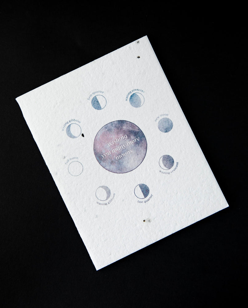 White greeting card featuring blue and purple watercolour illustrations of the moon's phases. The card reads "wishing you many more moons"