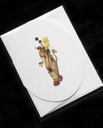 White oval greeting card with vintage-style illustration of a hand holding an apple with botanicals and spiders surrounding it.
