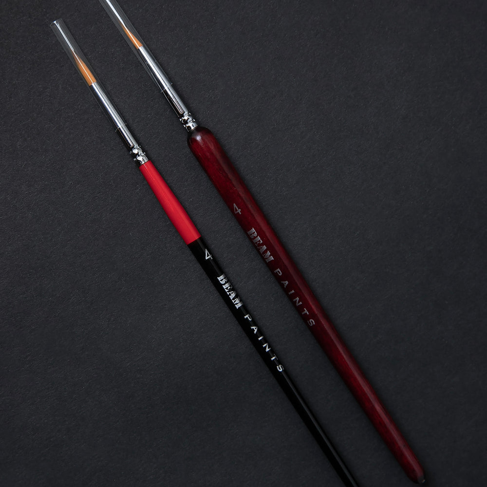 Two paintbrushes sitting side by side on a black background. One has a thin red and black handle, the other has a slightly thicker wooden handle stained red.