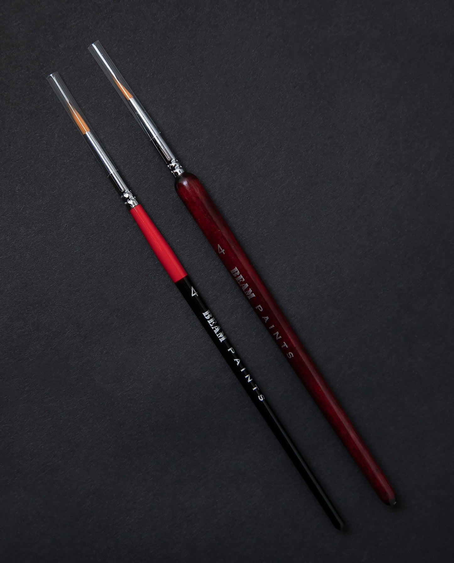Two paintbrushes sitting side by side on a black background. One has a thin red and black handle, the other has a slightly thicker wooden handle stained red.