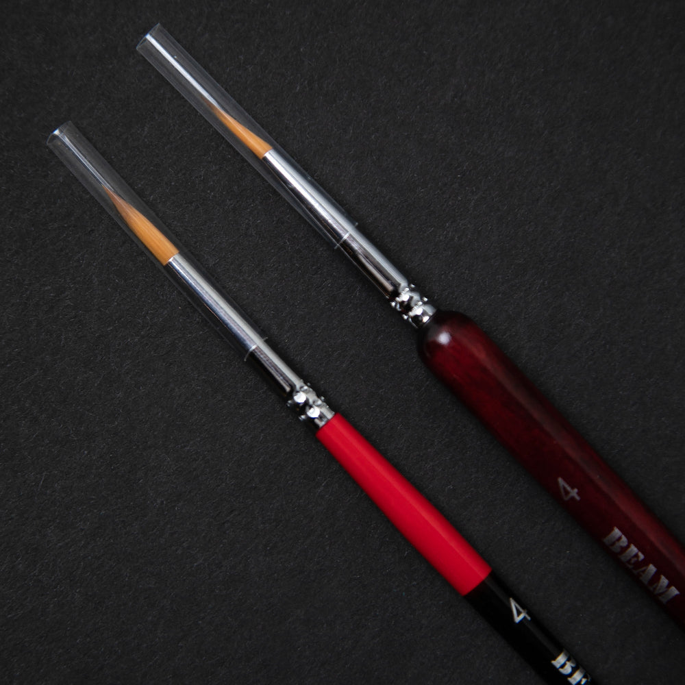 Close up on nylon bristles of two paintbrushes a black background. One has a thin red and black handle, the other has a slightly thicker wooden handle stained red.