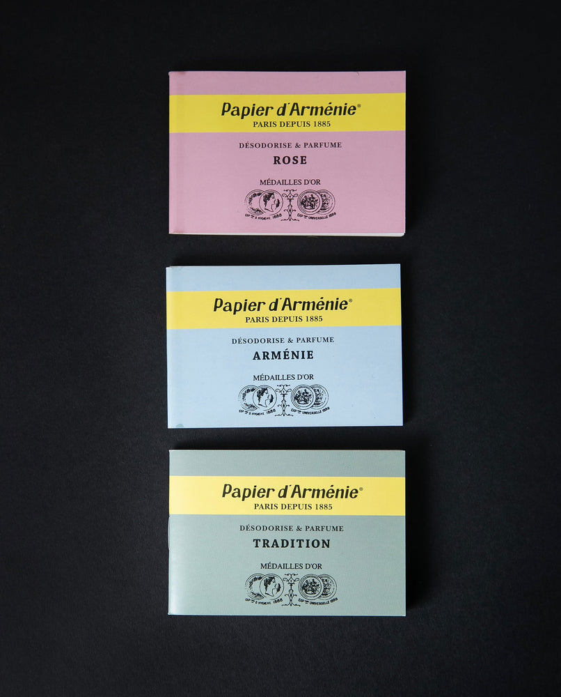 One of each of Papier d'Arménie's incense paper booklets on a black background. One is pink, one is baby blue, the other is sage green.