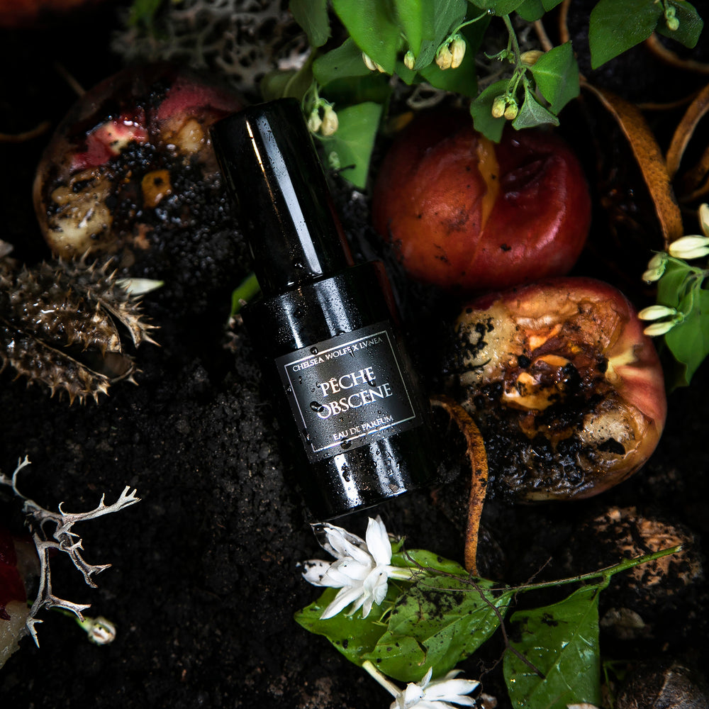 30ml black glass bottle of LVNEA best-selling natural perfume Peche Obscene lying on dirt surrounded by ripe peaches, jasmine, and botanicals