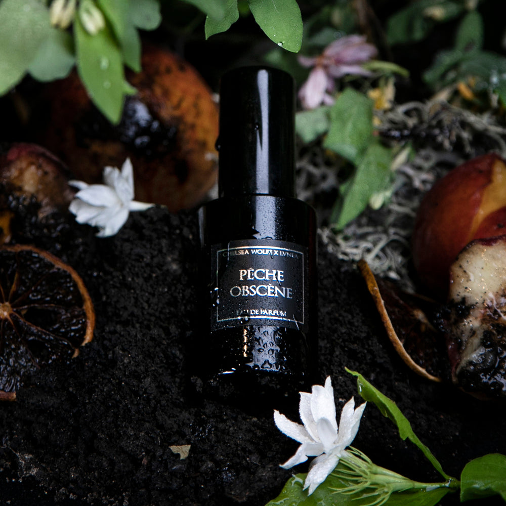 30ml black glass bottle of LVNEA best-selling natural perfume Peche Obscene lying on dirt surrounded by ripe peaches, jasmine, and botanicals
