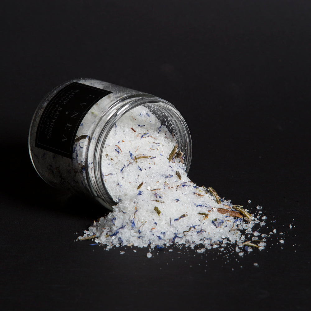 LVNEA'S River Bathing bath salts spilling out of a tipped over 16 ounce clear glass jar. There are blue cornflower petals, labrador tea buds, and usnea in visible in the salt mix.