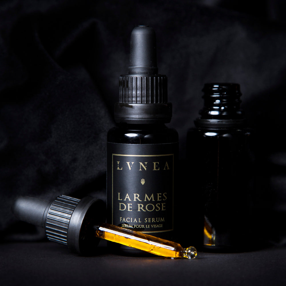 20ml black glass bottle with a black label reading "Larmes de Rose Facial Serum" in gold. There is a black unlabelled bottle next to it with its top removed, revealing a dropper filled with golden liquid.
